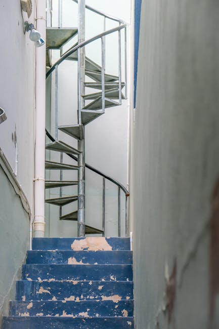 A narrow indoor staircase with blue painted steps, which are chipped and worn, leading upwards to a small landing with a spiral metal staircase adjacent to it. The spiral staircase has a silver metal 