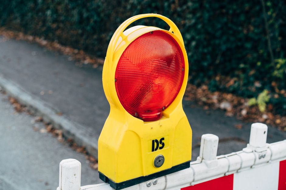 A yellow warning light atop a temporary road barrier, positioned on a pavement adjacent to a street with a curb and a white plastic barrier. The warning light has a large, circular red lens and is mou